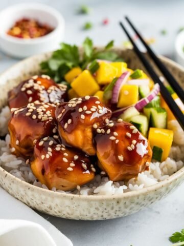 Sticky Teriyaki Salmon Bites Recipe served over fluffy white rice, drizzled with a glossy teriyaki glaze, and sprinkled with sesame seeds. Paired with a vibrant mango salsa featuring diced mango, cucumber, and red onion. A delicious and healthy seafood meal with bold flavors.