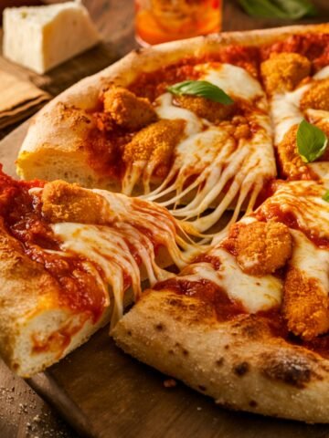 A homemade Chicken Parm Pizza with a golden-brown crust, crispy breaded chicken pieces, gooey melted mozzarella, and rich marinara sauce. A slice is being pulled, stretching the cheese. Fresh basil leaves add a pop of color, and the pizza sits on a rustic wooden board with a cozy, warm background.