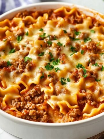 A homemade Hamburger Helper lasagna in a round white dish, topped with melted mozzarella, browned ground beef, and a sprinkle of fresh parsley. Curly lasagna noodles peek through a rich tomato meat sauce, with a blue-striped towel in the background on a white countertop.