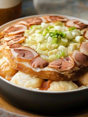 A close-up of a homemade Mille Feuille Nabe in a black pot, with layers of napa cabbage and slices of char siu (roast pork) on top. Fresh green onions garnish the dish, and a golden, clear broth surrounds the ingredients. The soft lighting gives the dish a warm, homemade look.