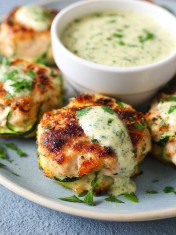 Plate of golden Chicken Zucchini Poppers, garnished with parsley, served with a creamy dipping sauce in a small white bowl. The dish has a fresh, homemade look with visible zucchini in the poppers.