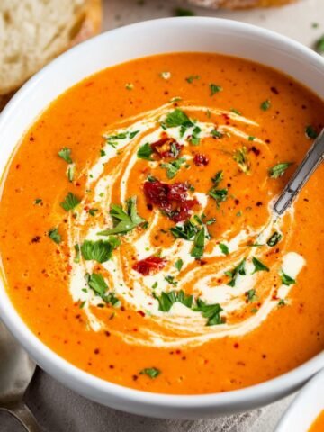 Homemade Roasted Red Pepper Gouda Soup, garnished with a swirl of cream, fresh parsley, and sun-dried tomatoes. A spoon is placed in the bowl, and a piece of bread is visible beside it.