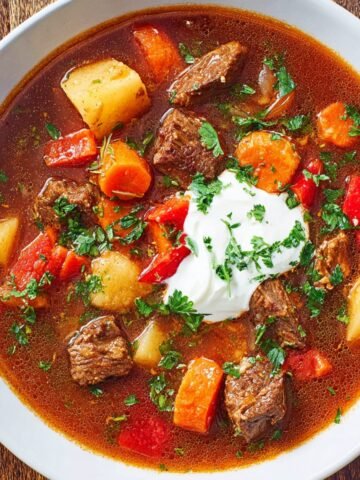 A hearty bowl of homemade Hungarian goulash filled with tender beef, potatoes, carrots, and red peppers, topped with a dollop of sour cream and fresh parsley.
