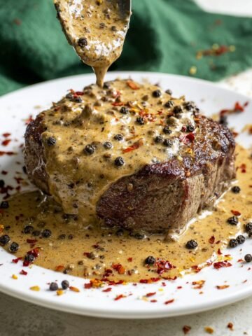 A juicy steak topped with creamy peppercorn sauce, sprinkled with black pepper and red chili flakes, resting on a white plate.