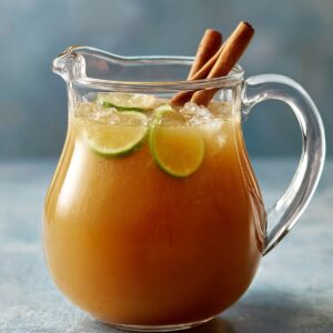 The Best Apple Cider Margarita Recipe 14 A glass pitcher filled with iced apple cider-style drink, garnished with lime slices and cinnamon sticks.