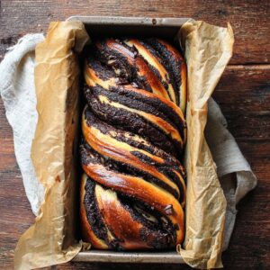 Best Babka Recipe | Chocolate Orange Swirl Bread 20 A freshly baked chocolate babka twisted loaf inside a parchment-lined loaf pan, glossy golden crust with dark chocolate layers visible, rustic wooden table background, cozy bakery aesthetic, overhead shot, natural light