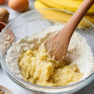 Easy Banana Bread Recipe - Ready in 70 Minutes | So Moist 16 A spatula is mixing chopped walnuts into banana bread batter in a large mixing bowl. The bananas in the background show the ingredients being prepared.