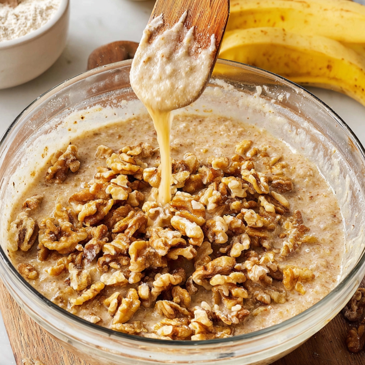 Easy Banana Bread Recipe - Ready in 70 Minutes | So Moist 12 "A hand pouring chopped walnuts into the banana bread batter in a large glass bowl. The bananas and other baking ingredients like flour are visible in the background."