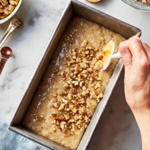 Easy Banana Bread Recipe - Ready in 70 Minutes | So Moist 17 "A hand is adding chopped walnuts to the top of the banana bread batter in a metal loaf pan. The background includes measuring spoons and a bowl of walnuts."