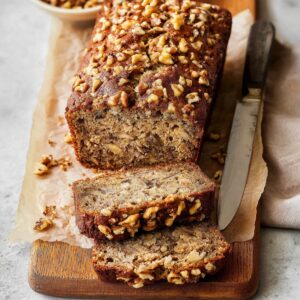 Easy Banana Bread Recipe - Ready in 70 Minutes | So Moist 18 "A freshly baked loaf of banana nut bread, topped with chopped walnuts. The bread is sliced to reveal its moist and nutty interior. The loaf is resting on parchment paper on a wooden cutting board, with a knife beside it."