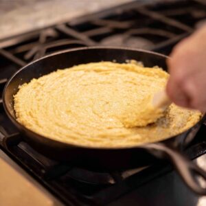 Cornbread Recipe: Easy 40-Minute Homemade Version 16 "A hand spreading cornbread batter evenly in a pan, preparing it for baking. The smooth batter is in a cast iron skillet."