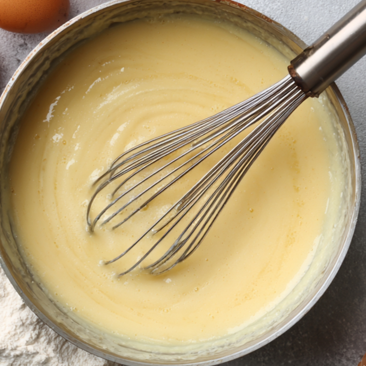 Delicious Crepe Recipe In 30 Minutes 10 A bowl of smooth crepe batter being whisked, surrounded by flour, eggs, and eggshells on a light countertop.