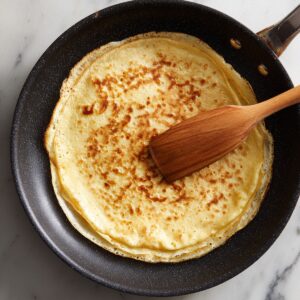 Delicious Crepe Recipe In 30 Minutes 14 A golden crepe cooking in a nonstick skillet, with a wooden spatula resting on top as it browns evenly.