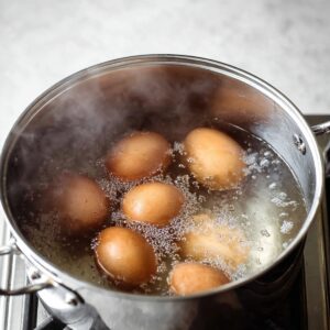 Deviled Eggs Recipe - Perfect in 35 Minutes or Less! 15 A stainless steel pot on the stove with several brown eggs submerged in boiling water, creating bubbles as the eggs cook. Steam rises from the water, indicating the eggs are boiling.