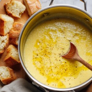 The Best French Garlic Soup Recipe 16 A pot of creamy garlic soup drizzled with olive oil and sprinkled with black pepper, accompanied by toasted bread cubes on a wooden board.