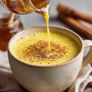 Delicious Golden Milk Recipe In 10 Minutes 16 A warm mug of golden milk topped with cinnamon, set on a wooden tray with a soft cloth, as honey is being poured in from a small glass jar.