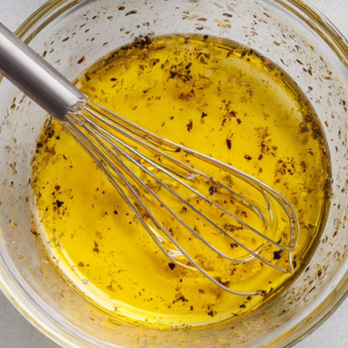 Healthy Greek Orzo Salad Recipe 11 Ingredients for Greek salad dressing laid out on a counter, including a bowl of whisked olive oil and herbs, uncooked orzo, dried oregano, and olive oil.