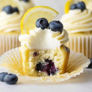 Lemon Blueberry Cupcakes Recipe (Moist & Easy!) 19 Lemon blueberry cupcake cut in half, showing a moist crumb filled with fresh blueberries, topped with white buttercream frosting, blueberries, and a lemon slice.
