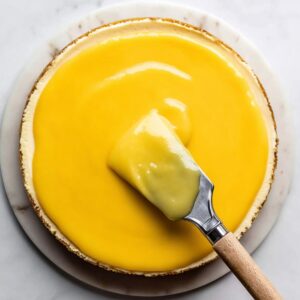 Best Lemon Cheesecake Recipe 18 Top-down view of lemon curd being spread across a baked cheesecake with a metal offset spatula