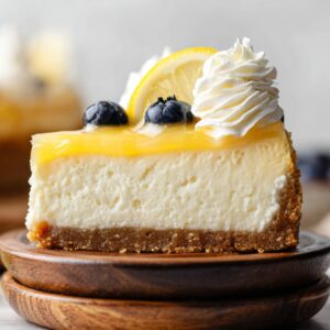 Best Lemon Cheesecake Recipe 19 Slice of lemon cheesecake topped with lemon curd, whipped cream, and blueberries on stacked wooden plates.