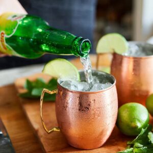 Best Moscow Mule Recipe in 5 Minutes | Easy Vodka Cocktail 16 Ginger beer being poured from a green bottle into copper mugs filled with ice for Moscow Mule cocktails.