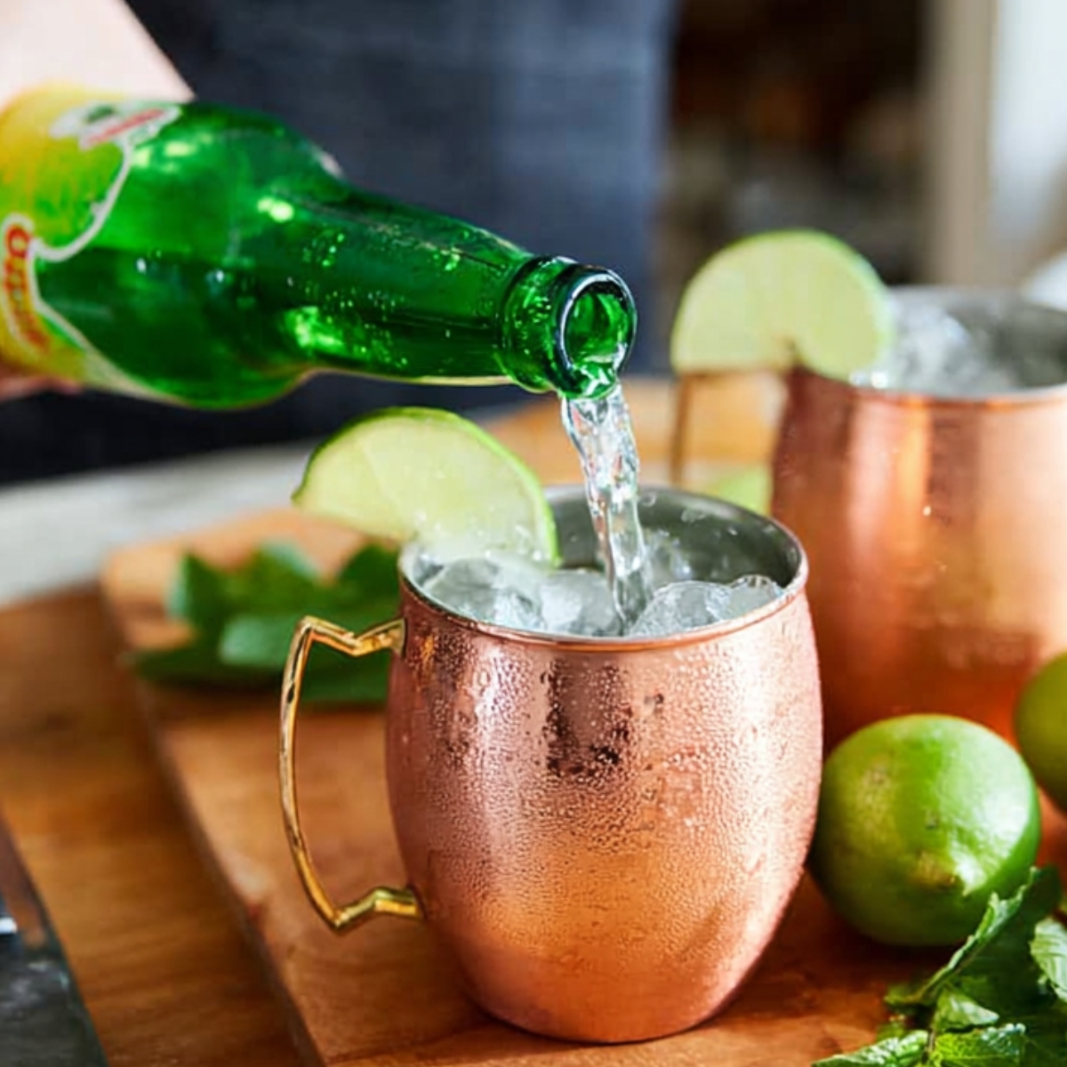 Best Moscow Mule Recipe in 5 Minutes | Easy Vodka Cocktail 12 Ginger beer being poured from a green bottle into copper mugs filled with ice for Moscow Mule cocktails.