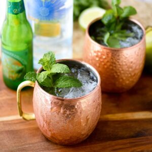 Best Moscow Mule Recipe in 5 Minutes | Easy Vodka Cocktail 17 Two copper mugs filled with ice-cold Moscow Mule cocktails garnished with fresh mint on a wooden board.