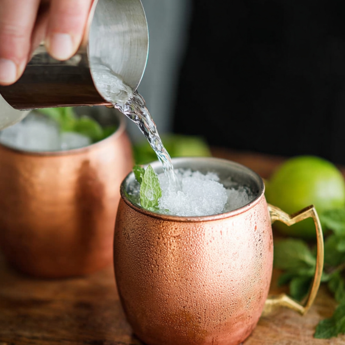 Best Moscow Mule Recipe in 5 Minutes | Easy Vodka Cocktail 10 Hand pouring clear vodka from a metal jigger into a copper mug filled with crushed ice for a Moscow Mule.