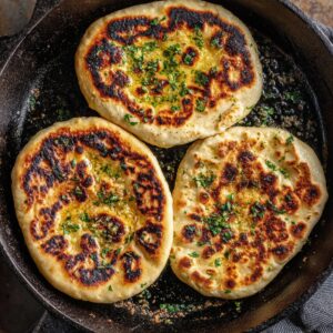 Healthy Naan Bread Recipe 16 Alt text: Garlic naan cooking in a cast-iron skillet, topped with melted butter and fresh herbs with golden char marks.