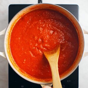 Nigerian Chicken Stew 15 A wooden spoon stirring thick Nigerian red pepper stew in a wide pot, smooth blended tomato and pepper sauce, rich red color, overhead view, clean modern stovetop, high-detail cooking process shot