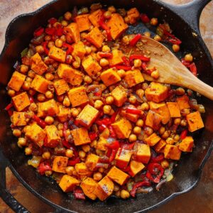 Easy Sweet Potato Hash Recipe | 5 Simple Ingredients 15 A cast iron skillet filled with spiced sweet potato cubes, chickpeas, red bell peppers, and onions being sautéed, wooden spoon stirring, rich warm tones, rustic kitchen surface, high-detail food photography, natural light.