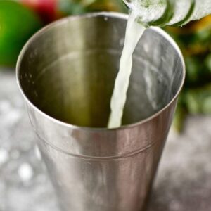 5-Ingredient White Christmas Mojito That's Ready in 10 Minutes! 15 "A person pouring a refreshing liquid, likely a cocktail, into a stainless steel shaker. The background shows a blurred kitchen setting with a few visible fruits like lime."