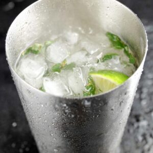 5-Ingredient White Christmas Mojito That's Ready in 10 Minutes! 14 "A stainless steel shaker filled with ice cubes and fresh mint leaves, with a slice of lime resting on top. The background is dark, enhancing the shiny texture of the shaker."