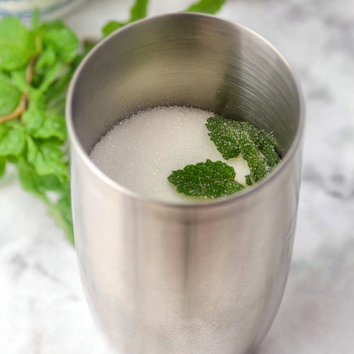 5-Ingredient White Christmas Mojito That's Ready in 10 Minutes! 12 "A close-up of a stainless steel cocktail shaker filled with sugar and fresh mint leaves, ready to be mixed. The shaker is set on a marble countertop with soft lighting."