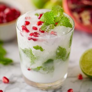 5-Ingredient White Christmas Mojito That's Ready in 10 Minutes! 16 "A refreshing glass of Mojito filled with ice, pomegranate seeds, mint leaves, and a slice of lime. The glass is set on a marble countertop with lime wedges and a pomegranate in the background."