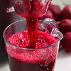 How to Make Beetroot Juice at Home - 2 Easy Ways 15 Close-up of fresh beet juice flowing from a juicer into a clear measuring cup, bright red color with frothy surface, whole beets visible in the background.