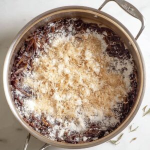 French Onion Soup Recipe - Rich & Easy in 1 Hour 15 Top-down view of caramelized onions in a stainless-steel pot sprinkled with flour during the soup-making process.