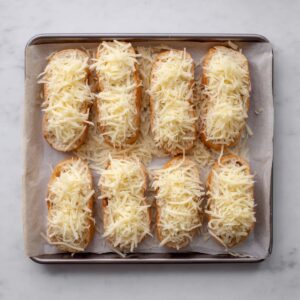 French Onion Soup Recipe - Rich & Easy in 1 Hour 16 Baking tray lined with parchment paper holding sliced baguette topped with shredded cheese, arranged evenly before baking.