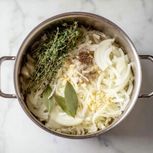 French Onion Soup Recipe - Rich & Easy in 1 Hour 14 Overhead view of sliced onions in a pot with fresh thyme, bay leaves, minced garlic, and seasonings before cooking.