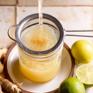 Homemade Ginger Shot Recipe | 5 Ingredients, 5 Minutes 16 Straining freshly pressed ginger juice into a jar with lime and ginger root beside, showcasing the natural ingredients