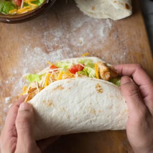 30-Minute Grilled Chicken Burrito - Crispy & Cheesy 15 Hands folding a large flour tortilla filled with grilled chicken, shredded cheese, lettuce, and tomatoes to form a chipotle ranch chicken burrito.