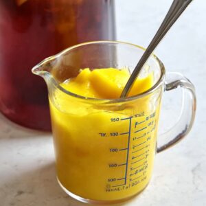 3-Ingredient Mango Iced Tea That's Better Than Starbucks! 15 "A measuring cup filled with mango puree, stirred with a spoon. In the background, a pitcher of iced tea is visible, showing that the puree is being added to the beverage."