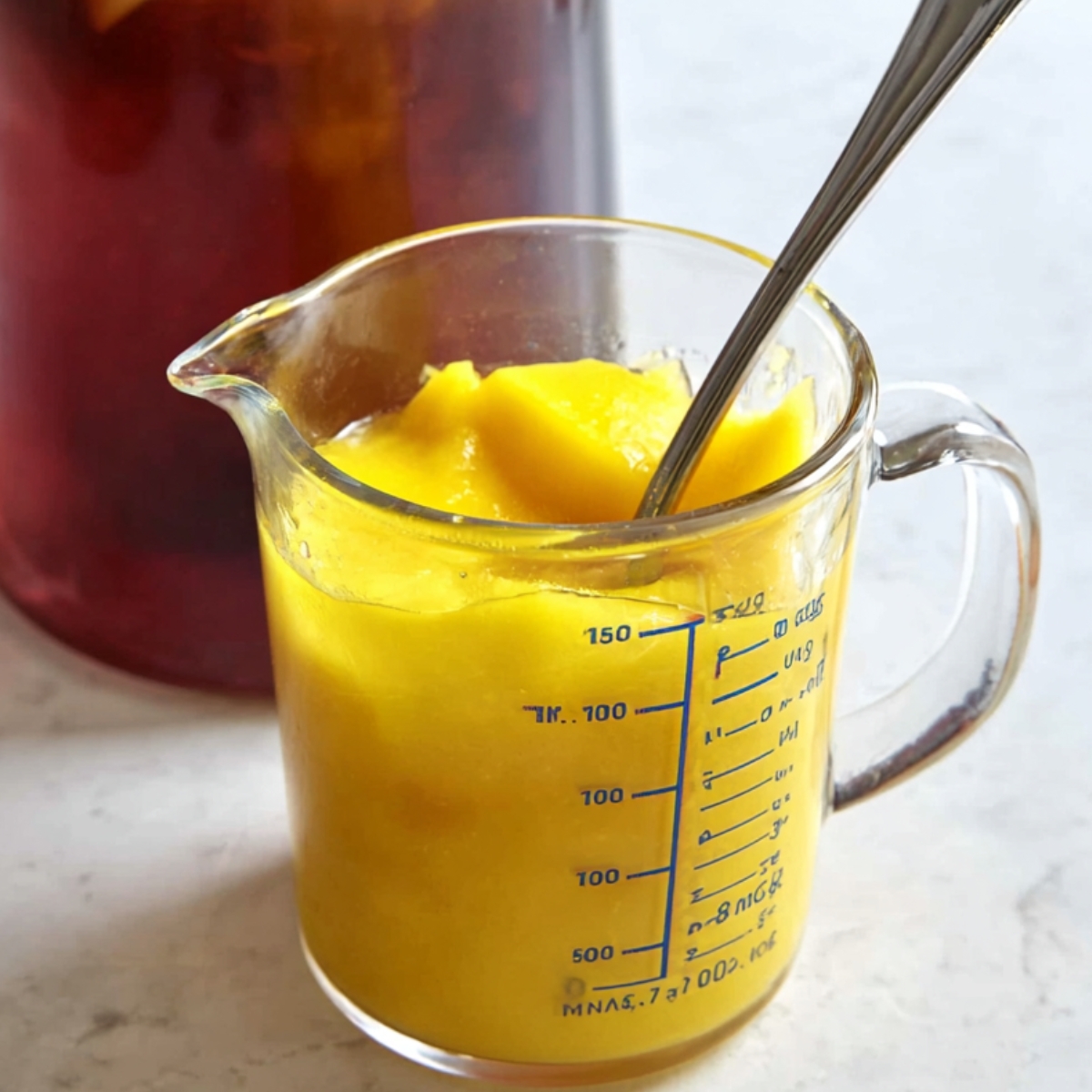 3-Ingredient Mango Iced Tea That's Better Than Starbucks! 11 "A measuring cup filled with mango puree, stirred with a spoon. In the background, a pitcher of iced tea is visible, showing that the puree is being added to the beverage."