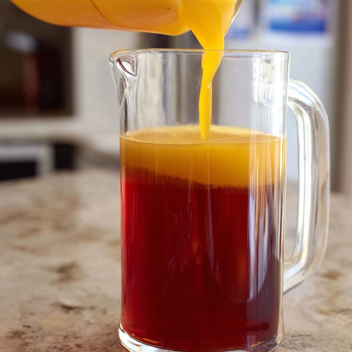 3-Ingredient Mango Iced Tea That's Better Than Starbucks! 12 "Mango juice being poured into a clear glass pitcher, mixing with red tea, creating a layered effect of orange and red. The background is blurred with soft lighting."