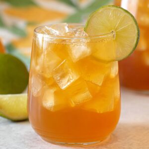 3-Ingredient Mango Iced Tea That's Better Than Starbucks! 17 "A refreshing glass of iced tea filled with ice cubes, garnished with a slice of lime. The background features a pitcher of iced tea and fresh limes."