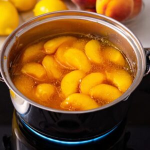 Peach Lemonade Recipe: Fresh, Easy & Ready in Minutes 15 A saucepan on the stove with peach slices cooking in a syrupy mixture. Lemons and fresh peaches are placed on a white surface next to the pan, while a jar of peach lemonade sits nearby."