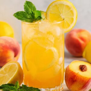 Peach Lemonade Recipe: Fresh, Easy & Ready in Minutes 17 A refreshing glass of peach lemonade filled with ice and garnished with lemon slices and mint leaves. Peaches and lemons are placed beside the glass on a white surface