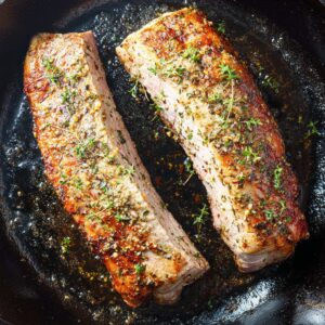 Best Pork Tenderloin Recipe | 30-Minute Dinner 15 Two pork tenderloins searing in a cast iron skillet, golden brown crust with herbs and spices, viewed from above.