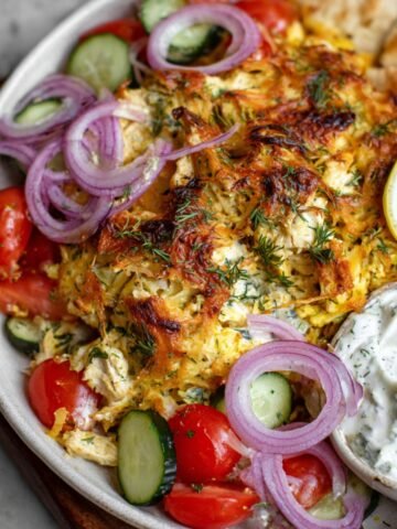 A close-up view of a Chicken Tzatziki Casserole served with a side of rice, garnished with onions, lemon wedges, and a creamy white sauce.
