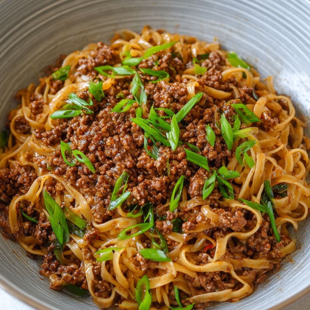 Mongolian ground beef noodles topped with sliced green onions, served in a ceramic bowl, ready to eat.